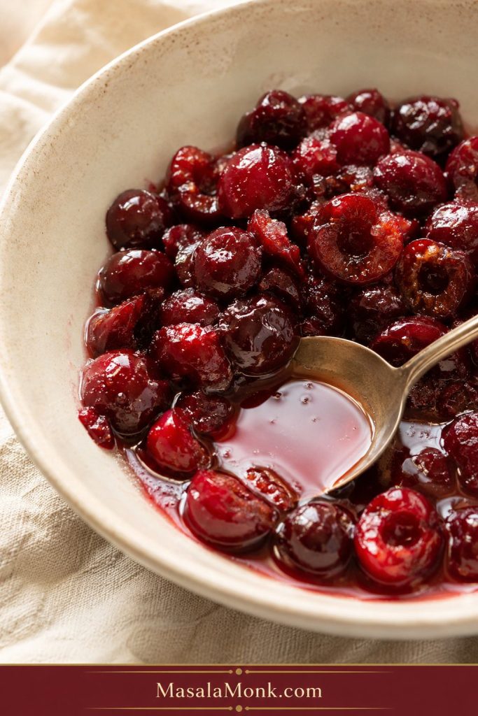 Pitted cherries mixed with sugar and lemon juice in a bowl with red syrup pooling around a spoon.