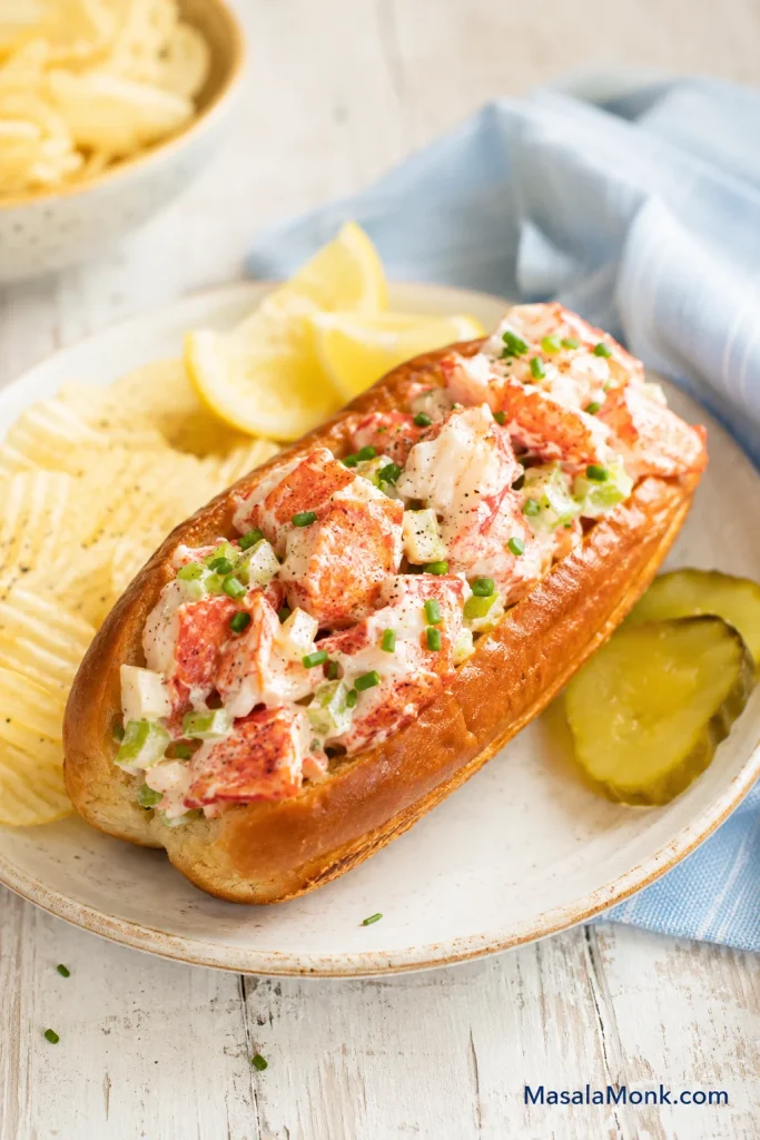 Finished Maine-style lobster roll with cold lightly dressed lobster, diced celery, chives, potato chips, pickles, lemon wedges, and a toasted bun.