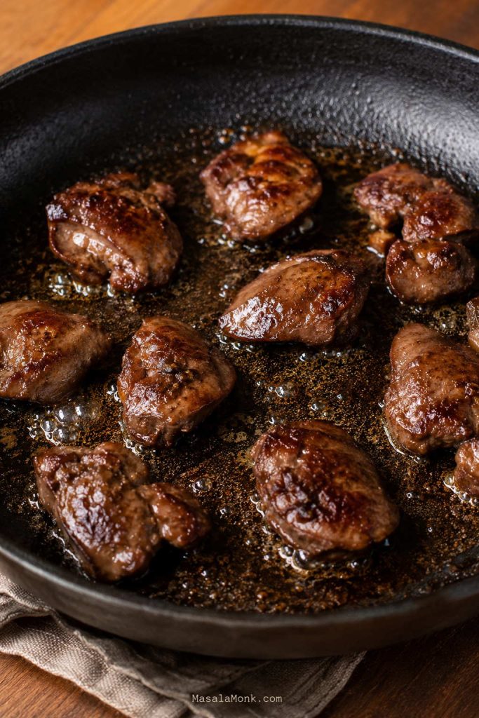Chicken livers searing in one layer in a skillet with space between the pieces and oil bubbling lightly.