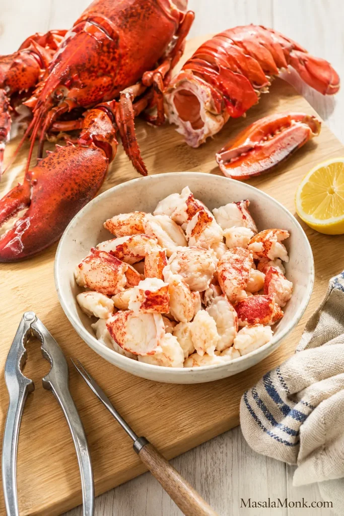 Picked lobster meat in a bowl beside cooked lobster shells, cracked claws, a tail shell, lobster crackers, lemon, and a kitchen towel.