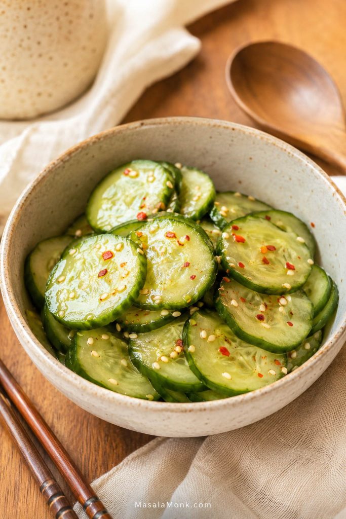 Ceramic bowl of sliced cucumber tossed with sesame seeds, chili flakes, and a glossy vinegar-style dressing.