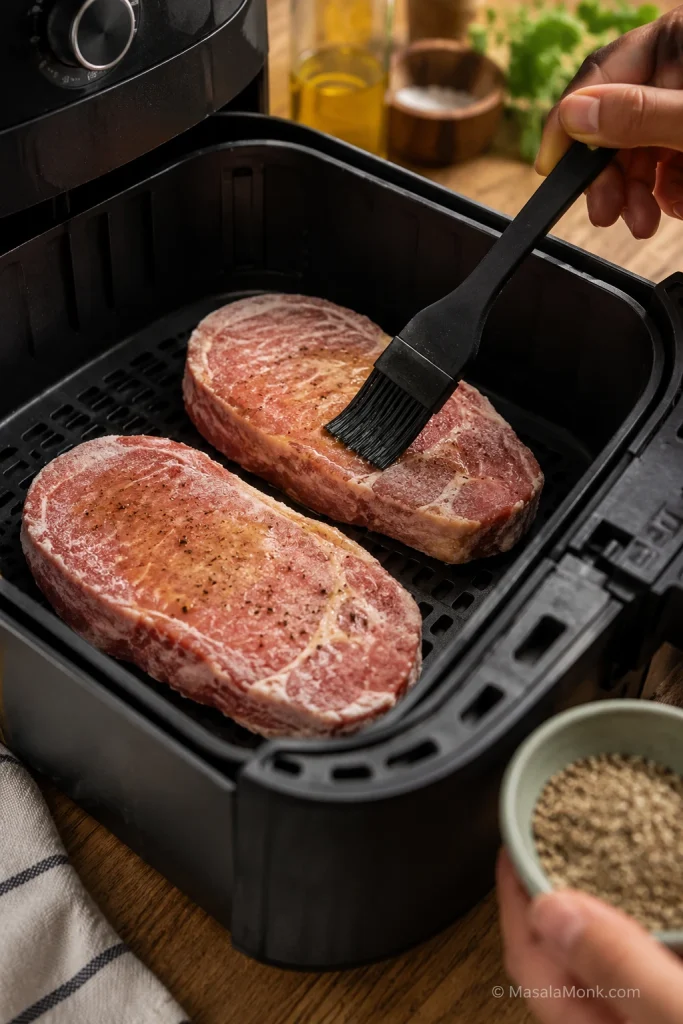 Frozen pork chops being brushed with oil and seasoning in an air fryer basket.