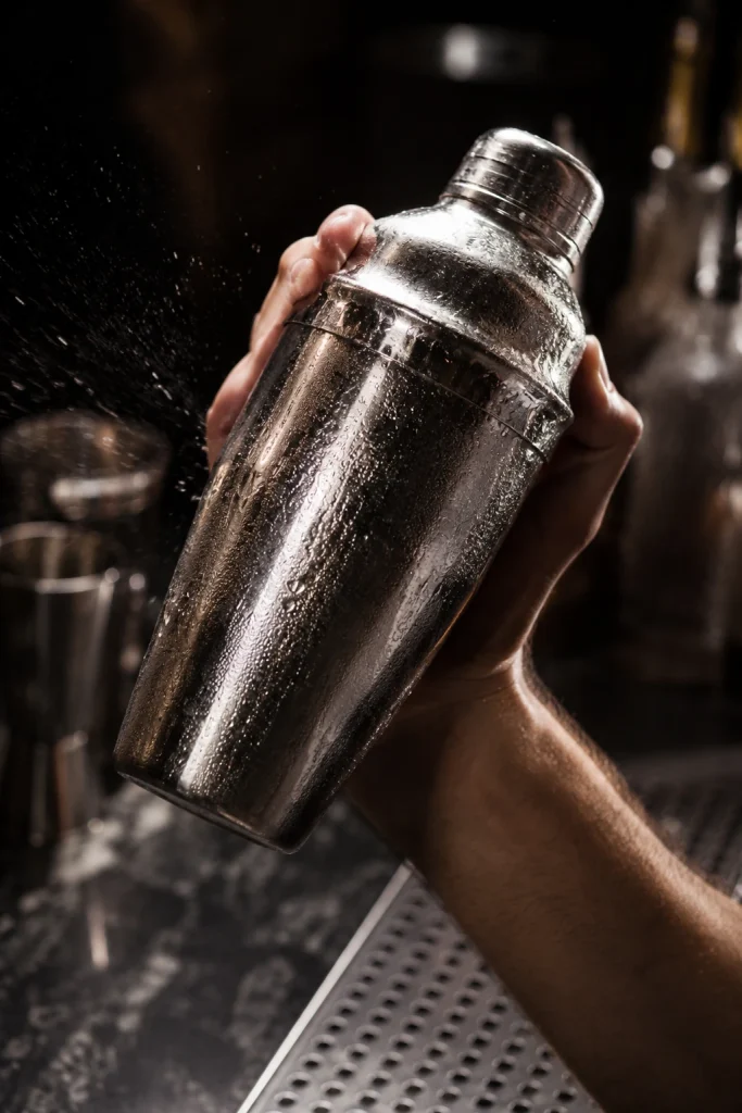 A hand shaking a chilled metal cocktail shaker covered in condensation in a dark bar setting.