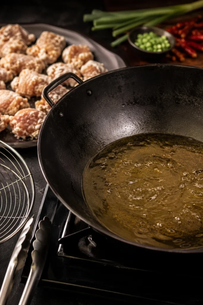 Dark wok with a shallow layer of oil on a stovetop, with coated chicken pieces waiting nearby on a tray and frying tools at the side.