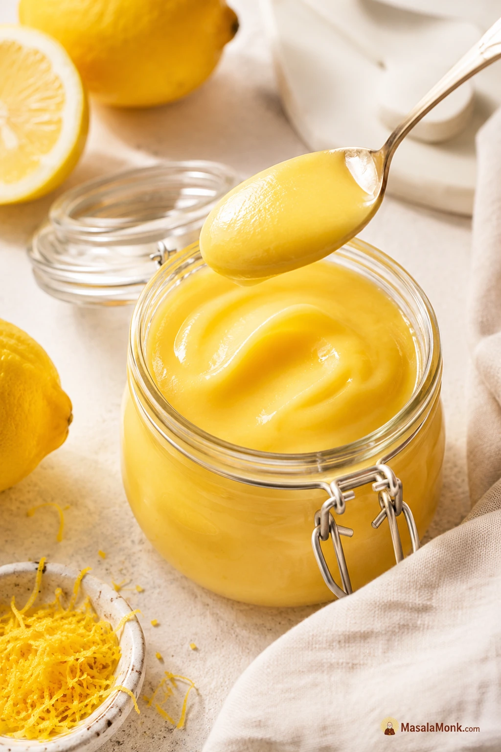 A glass jar of glossy homemade lemon curd with fresh lemons, lemon zest, and a spoonful lifted above the jar.