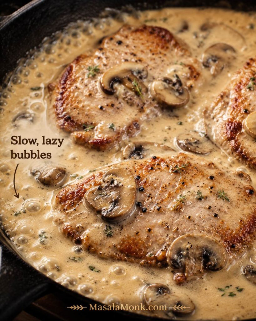 Pork chops simmering gently in creamy mushroom gravy with small bubbles around the edges and text reading slow lazy bubbles.