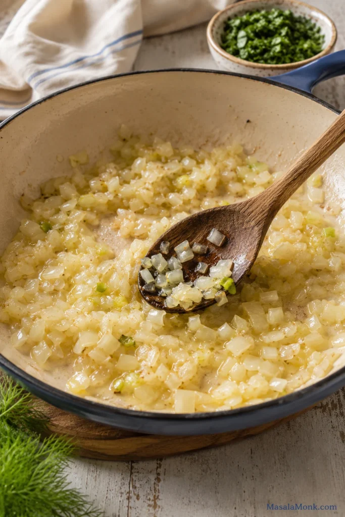 Softened onion, scallion, and garlic cooking in a skillet with a wooden spoon.