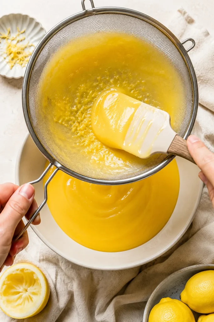 Warm lemon curd being pushed through a fine mesh sieve into a bowl, with small bits caught in the sieve.