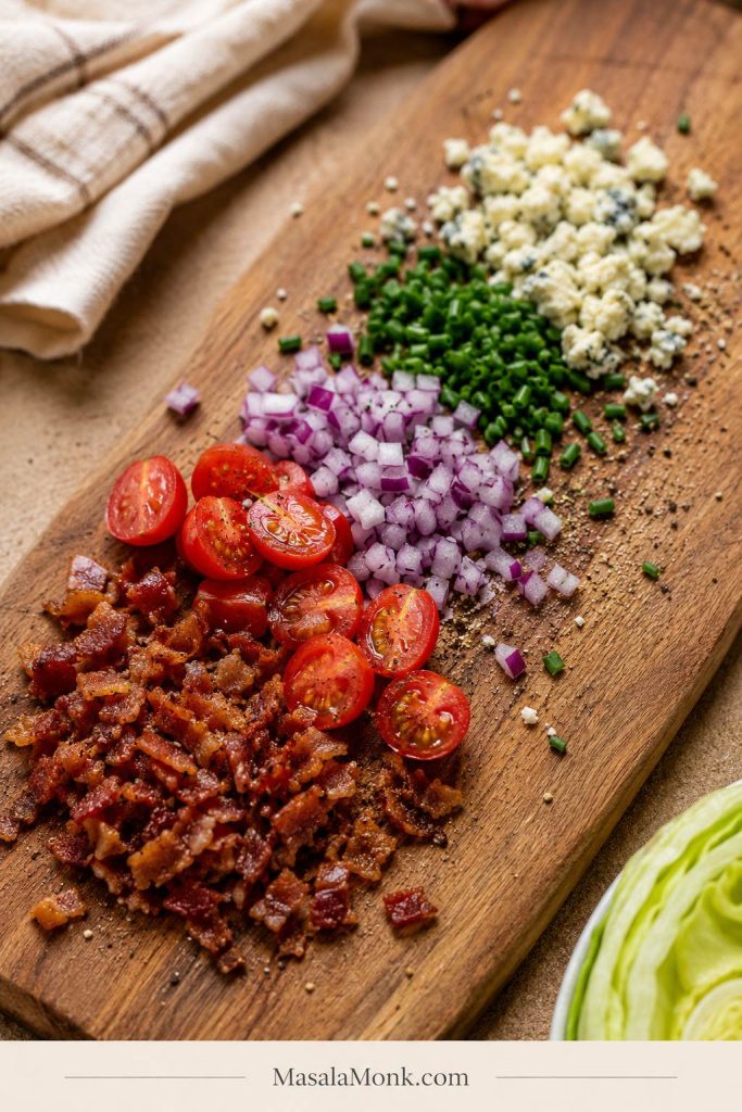 Small bacon bits, halved cherry tomatoes, diced red onion, chopped chives, blue cheese crumbles, and black pepper arranged on a wooden board.