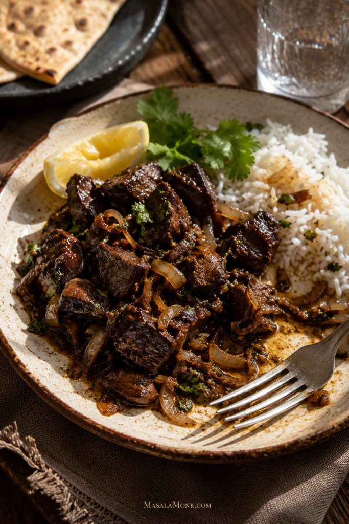 Chicken liver with onions served over rice with lemon, herbs, roti, and salad in the background.