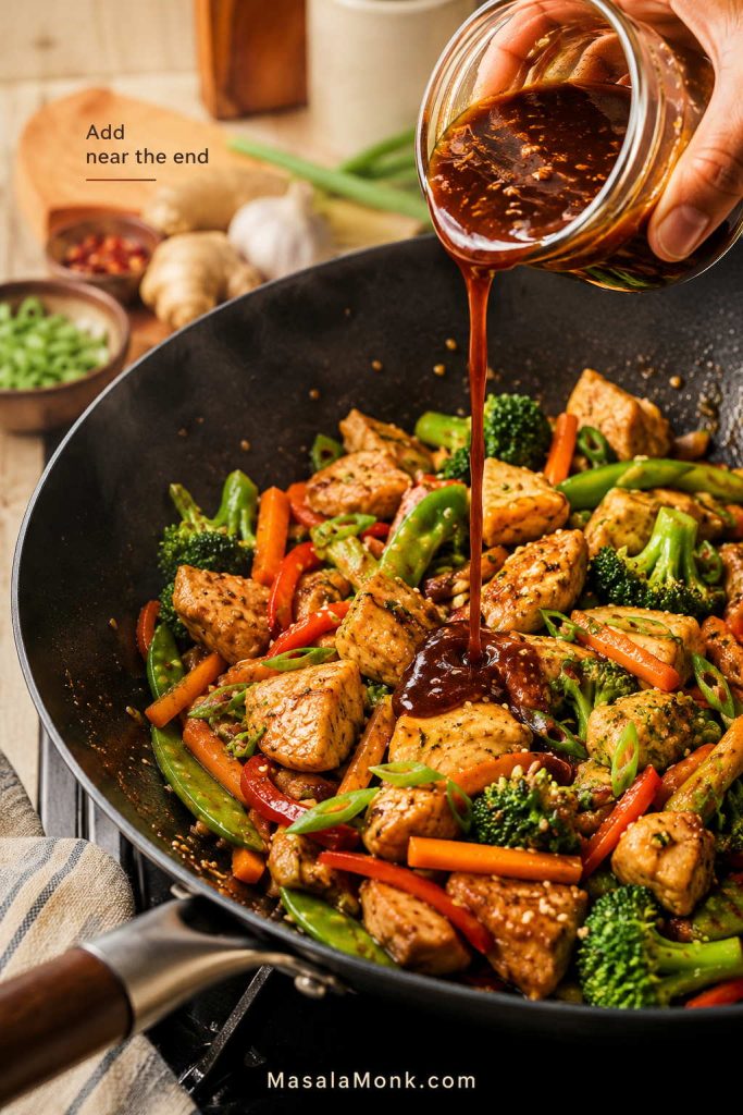 Brown stir fry sauce being poured from a jar into a wok of mostly cooked chicken, broccoli, carrots, peppers, snap peas, and scallions.