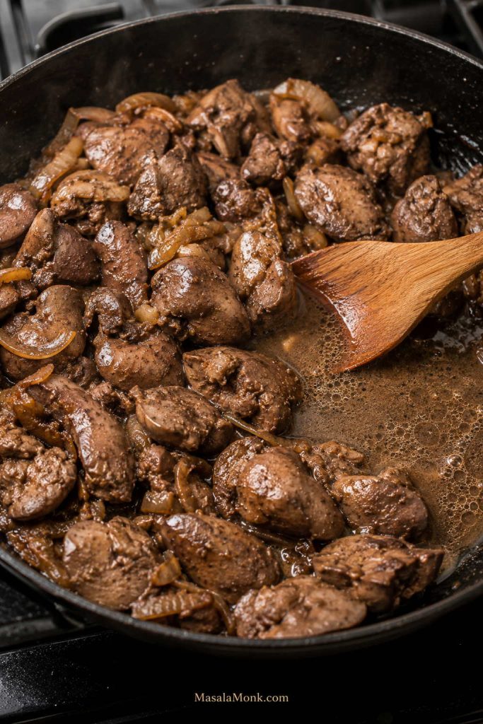 Crowded skillet of chicken livers and onions with visible liquid pooling beside a wooden spoon.
