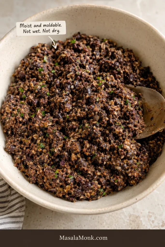 Bowl of chunky black bean burger mixture with visible black beans, oats, herbs, onion, and a spoon, labeled moist and moldable.