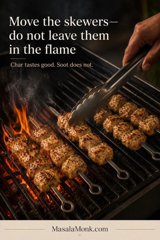 Tongs move chicken souvlaki skewers away from an active flare-up toward a cooler section of the grill.