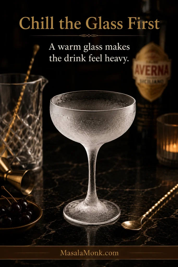 An empty frosted coupe glass with condensation on a dark bar surface beside a mixing glass, bar spoon, cherries, and an Averna bottle.