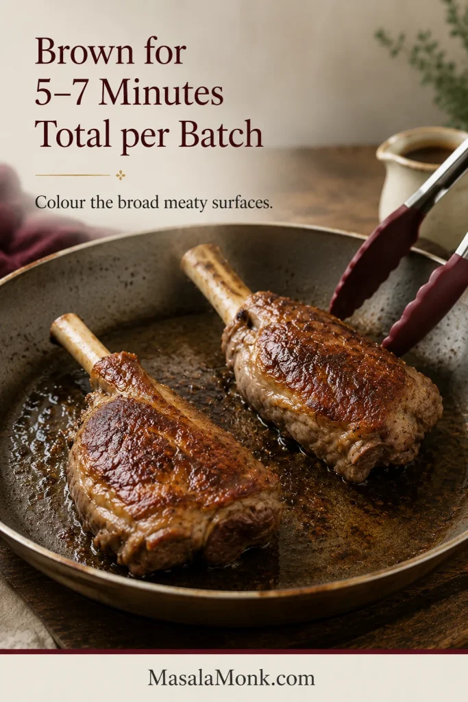 Two lamb shanks browning in a wide pan as silicone-tipped tongs turn one deeply browned meaty side.