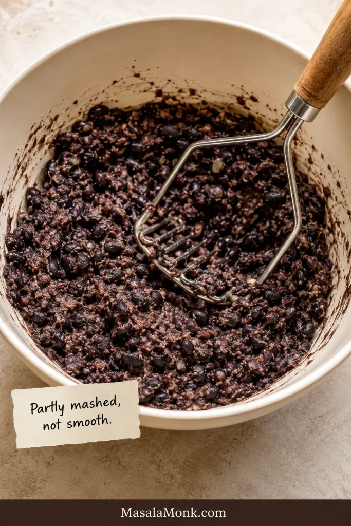 Bowl of partly mashed black beans with a fork or masher, showing some mashed beans and some whole bean pieces.
