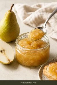 Open jar of pale-gold pear jam beside toast, fresh pears and a spoon on a softly lit breakfast table.