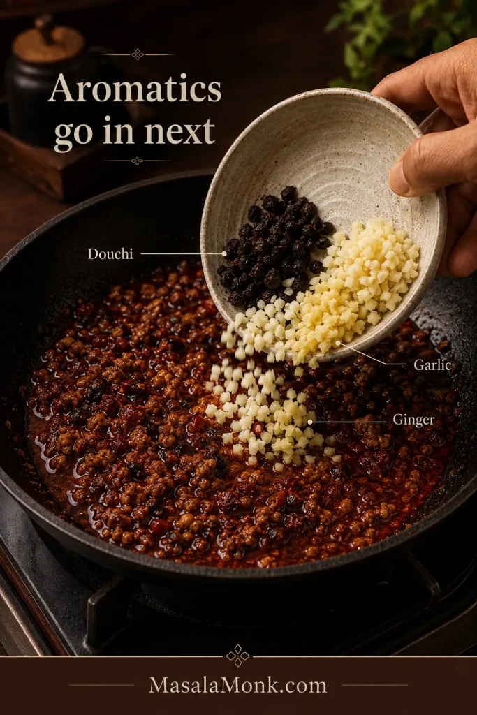 Douchi beans, minced garlic and minced ginger being tipped into a dark wok containing browned pork and a brick-red doubanjiang base.