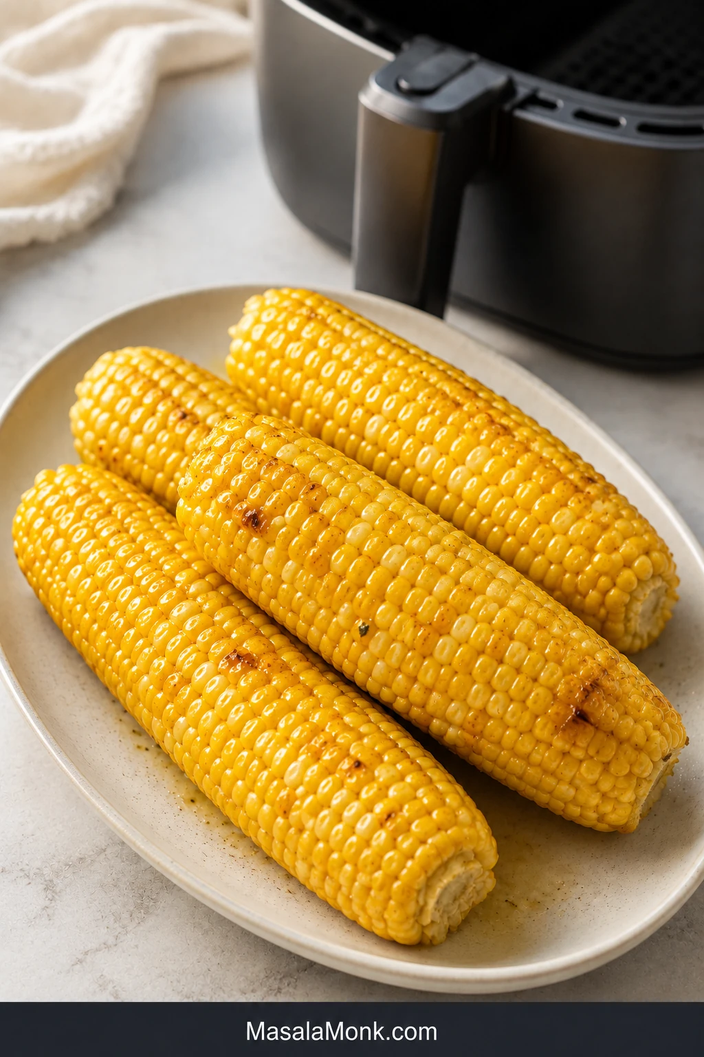 Three buttered ears of air-fried corn on a cream platter, with plump golden kernels, scattered roasted patches and an air fryer behind them.