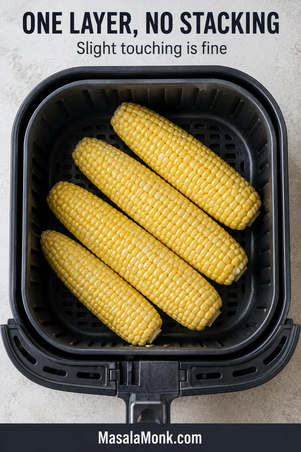 Four raw corn ears arranged diagonally in one layer inside a black air-fryer basket, with small spaces between them.