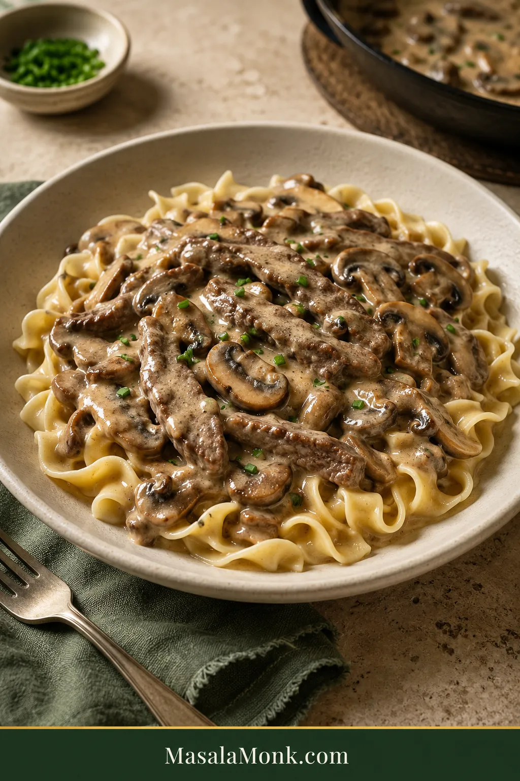 Beef Stroganoff over wide egg noodles with thin browned beef strips, sliced cremini mushrooms, glossy tan sauce and a small scattering of chopped chives.