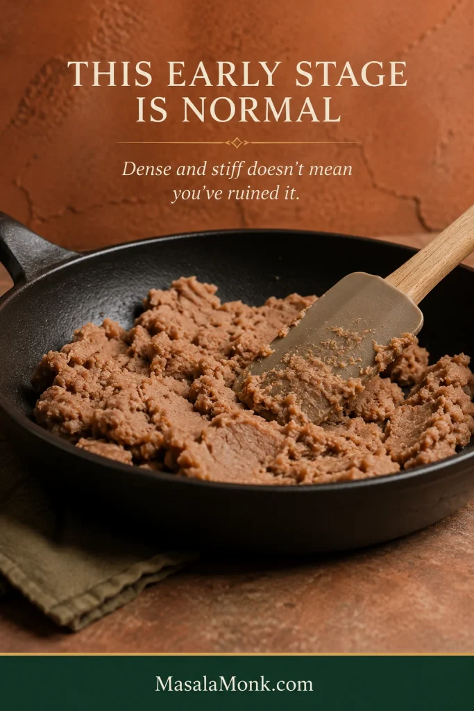 Dense canned refried beans in a dark skillet being pressed and folded with a spatula before they loosen.