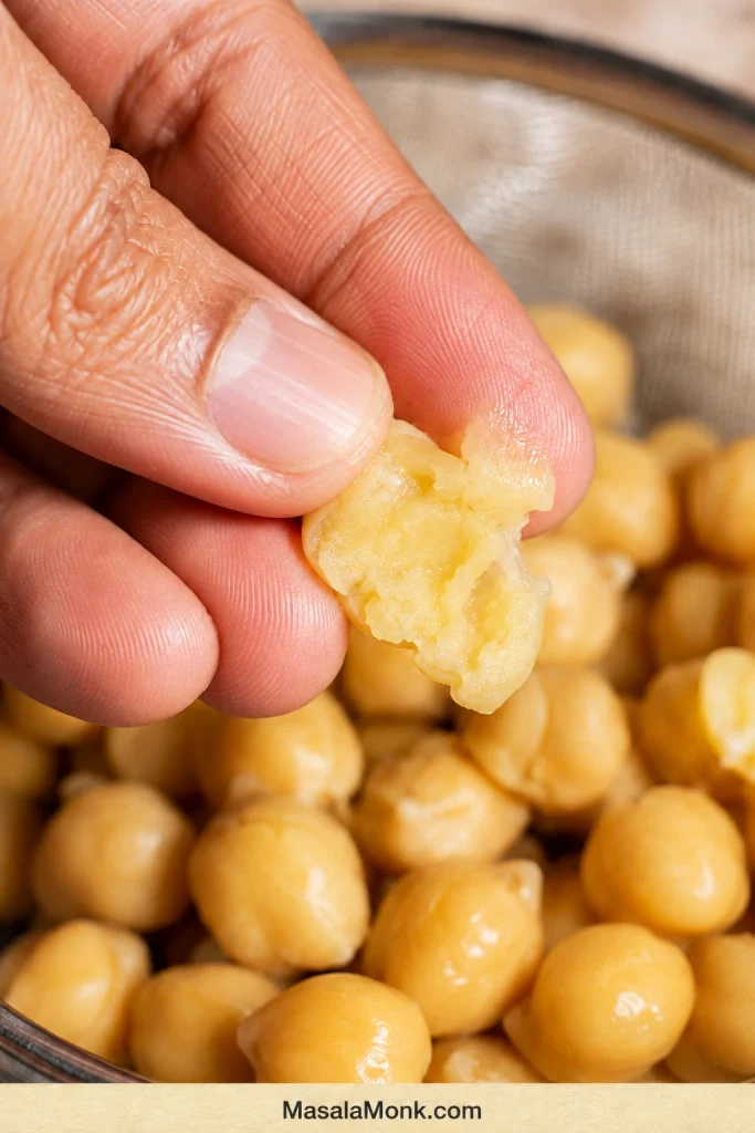 Thumb and forefinger pressing a soft cooked chickpea above a sieve filled with drained chickpeas.
