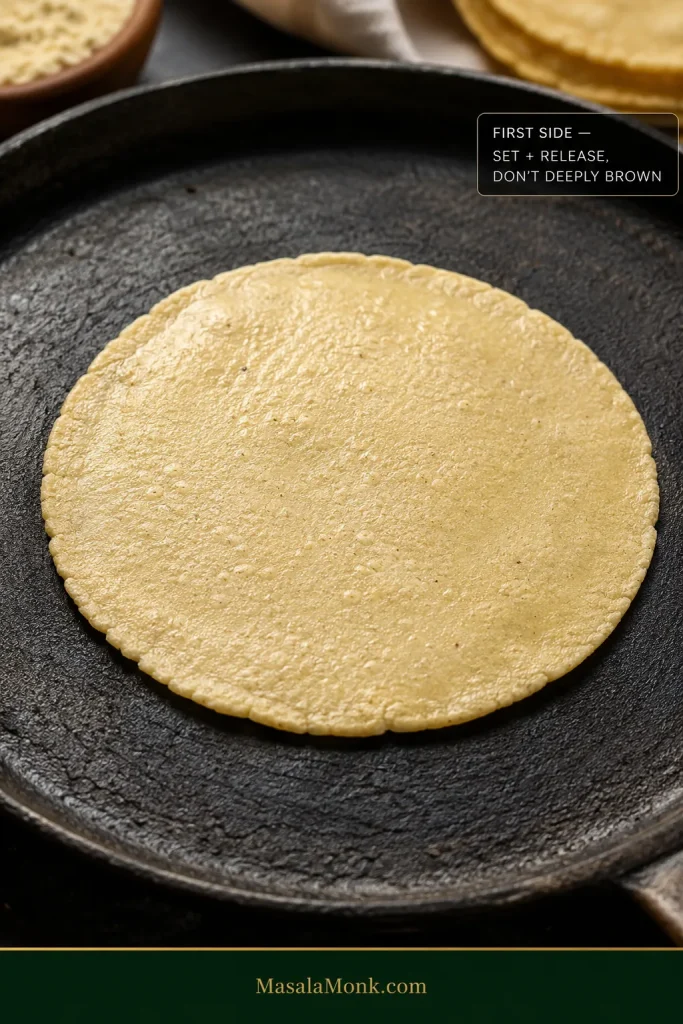 Pale corn tortilla cooking on its first side with little browning, a fading wet sheen, and edges beginning to release from the skillet.