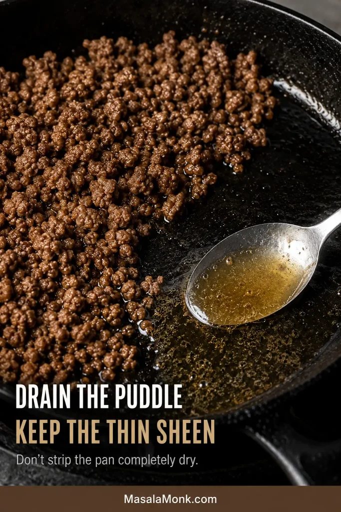Spoon removing pooled rendered fat from browned ground beef while leaving a thin sheen in the skillet.