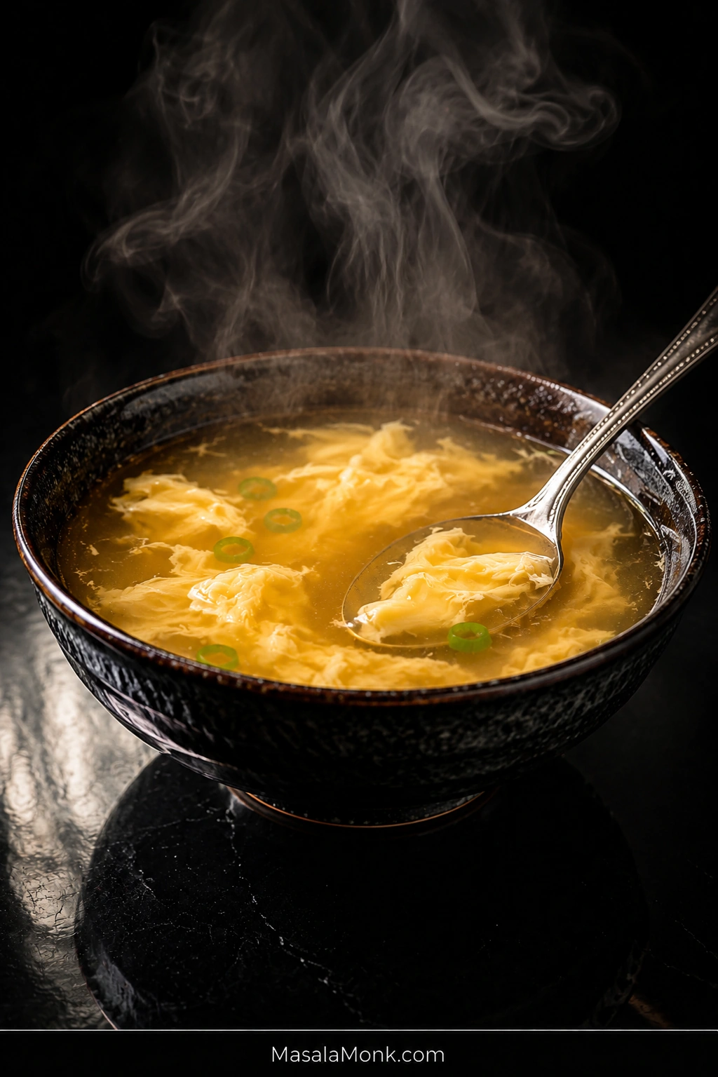 Steaming black bowl of golden egg drop soup with broad pale-yellow egg ribbons, sliced scallions, and a metal soup spoon on a dark reflective table.