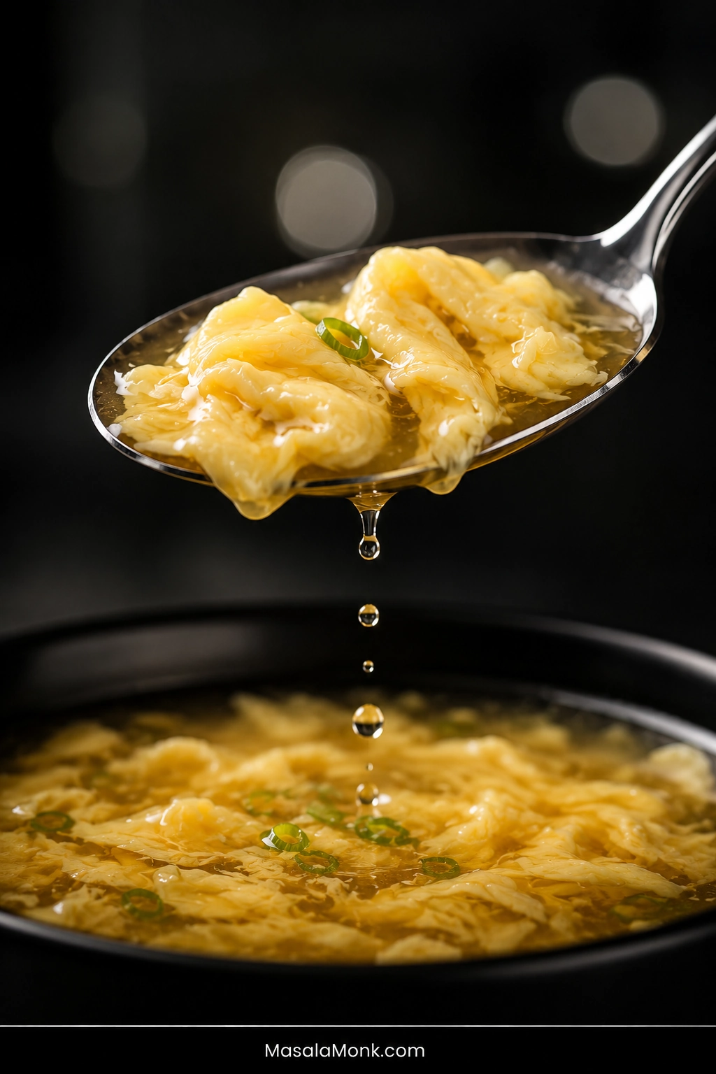 Close-up metal spoon lifting several soft folded egg ribbons and golden broth above a bowl of finished egg drop soup, with droplets falling from the spoon.