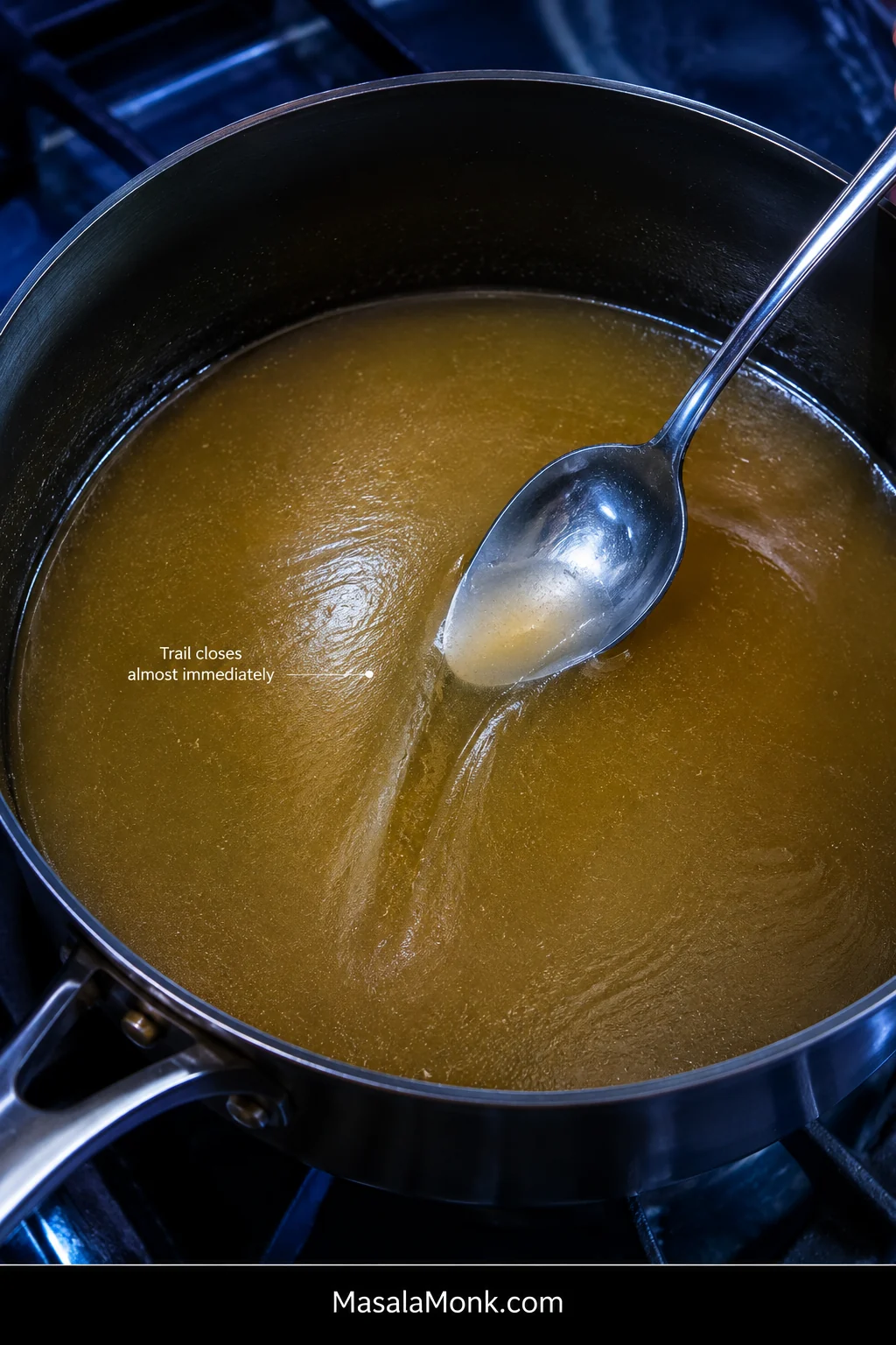 Metal spoon dragged through lightly thickened golden broth in a stainless saucepan, leaving a shallow trail that begins closing behind the spoon.