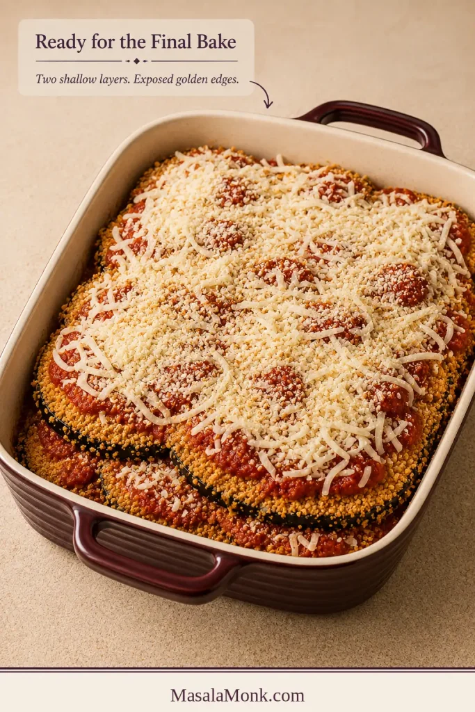 Unbaked layered Eggplant Parmesan with marinara, shredded cheese and exposed breaded edges in a burgundy casserole.