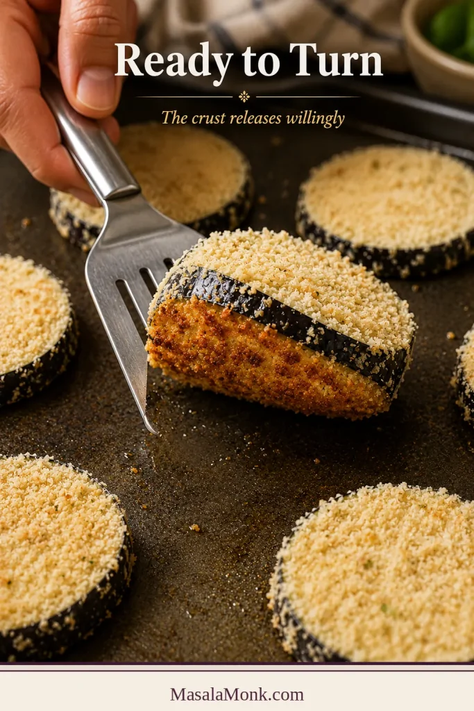 Spatula lifting a breaded eggplant round to reveal a golden underside while the upper surface remains pale.
