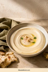 Wide ivory bowl of smooth hummus with olive oil, chickpeas, parsley and red spice, surrounded by warm pita on a pale stone table.