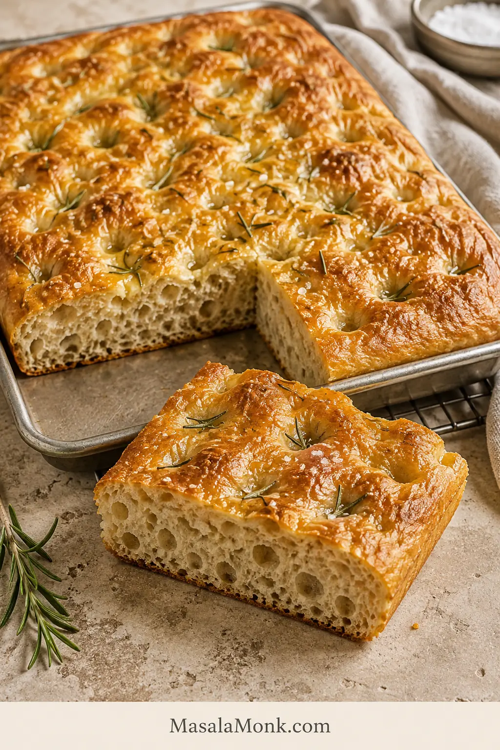 Golden rosemary focaccia with a cut square in front showing an airy crumb and thin browned bottom crust.