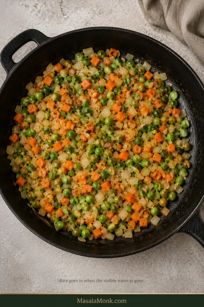 Peas, diced carrots and onion cooking in a skillet with steam rising but no visible puddle of liquid on the exposed pan surface.