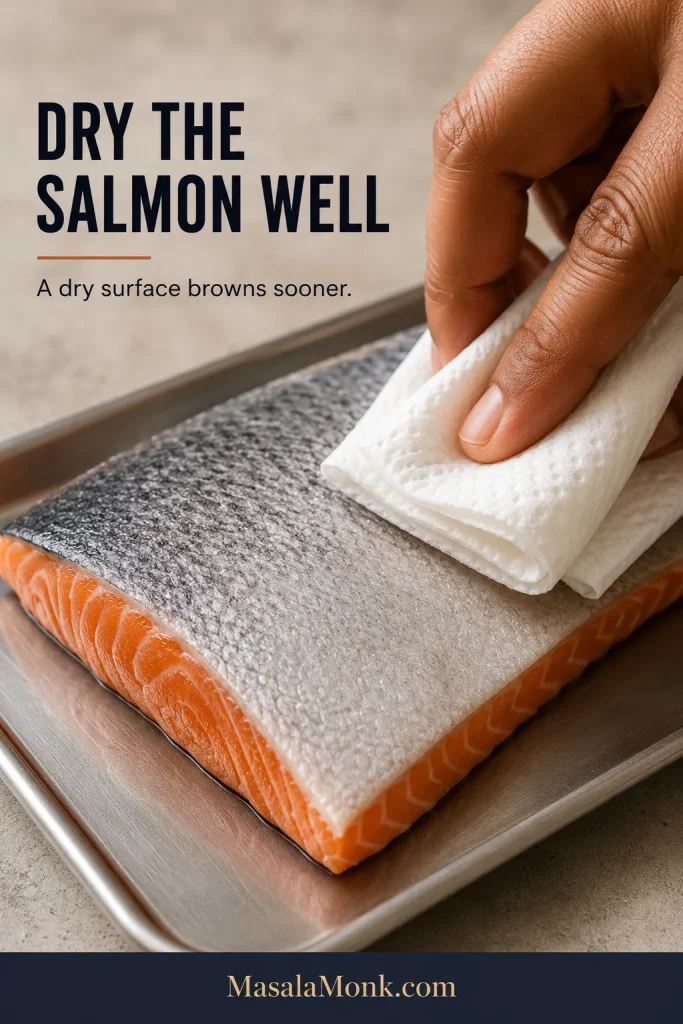 Hand gently blotting the skin of a raw salmon fillet with folded white paper towel on a metal tray.
