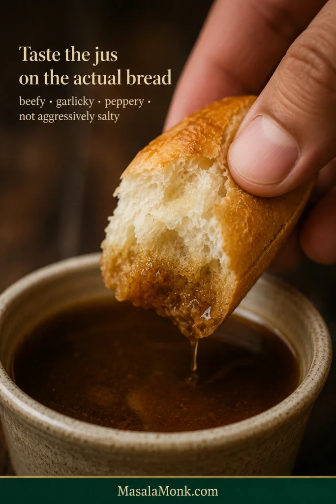 Hand dipping a torn piece of French roll into a bowl of dark brown Italian beef jus as the crumb absorbs the liquid.