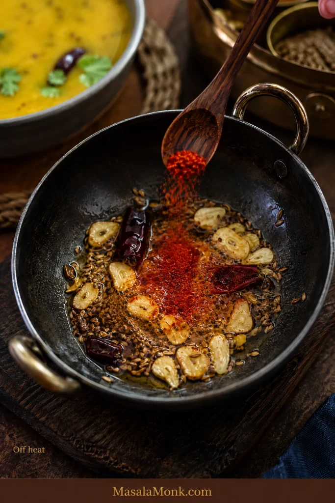 Kashmiri chilli powder being added to hot ghee with cumin, golden garlic and dried chilli away from the burner.