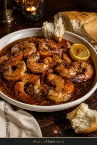 New Orleans BBQ shrimp in a shallow ivory bowl with glossy dark peppery sauce, lemon and torn French bread.