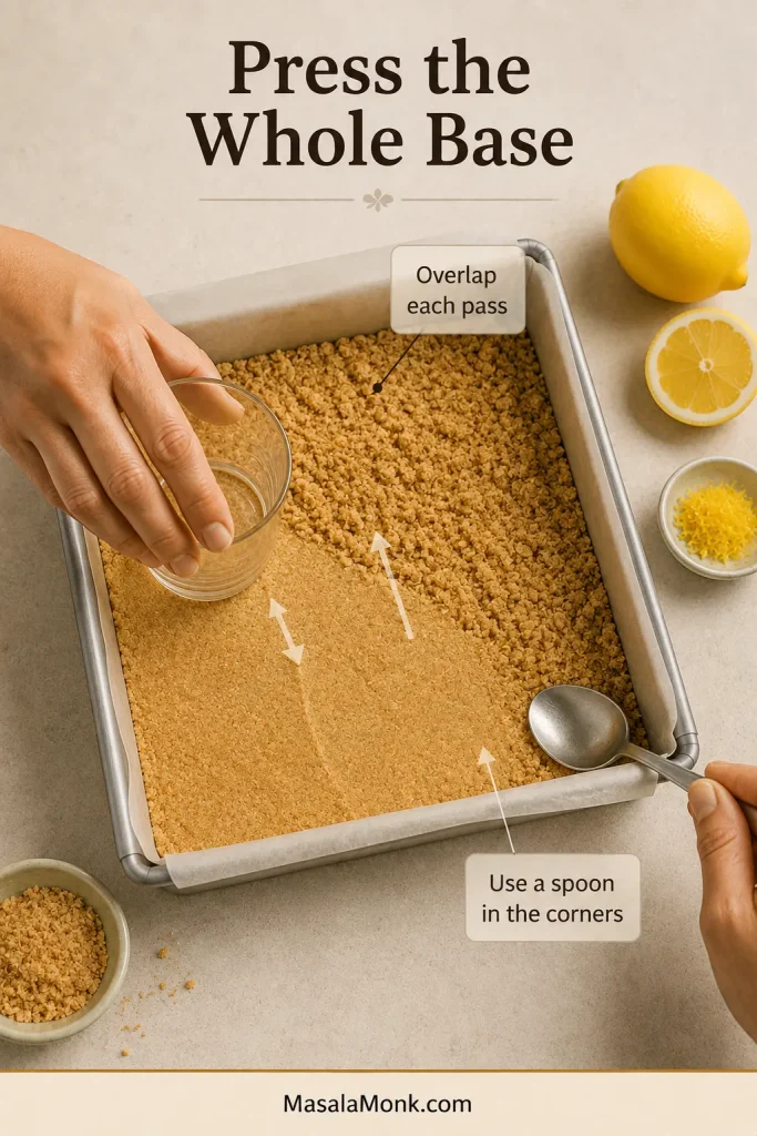 A flat-bottomed glass compacts biscuit crumbs across a lined pan while a spoon presses firmly into the corners.