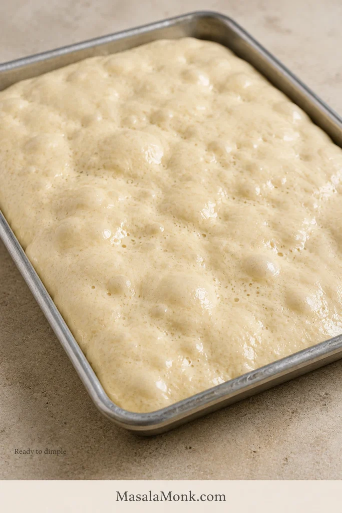 Close view of fully proofed focaccia dough filling a metal pan with a relaxed surface and many visible bubbles.