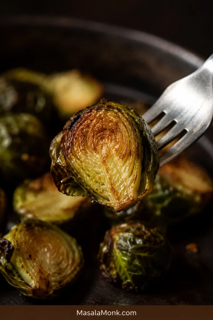 Fork holding a roasted Brussels sprout half with a deeply caramelized exterior, curled crisp leaf and tender green center.