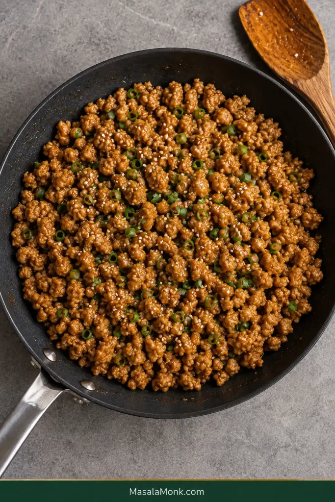 Overhead skillet of browned sesame ground chicken coated in a thin amber glaze with scallions and sesame seeds.