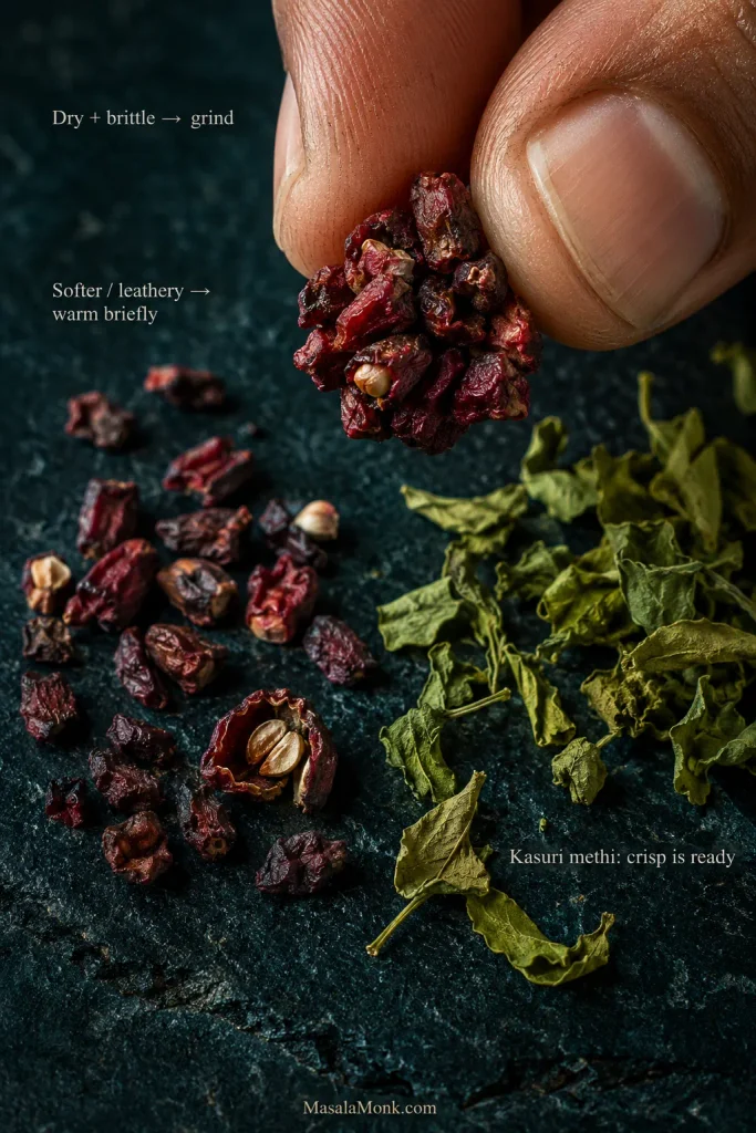 Close view of dried pomegranate arils held between fingers beside loose anardana and crisp dried kasuri methi leaves.
