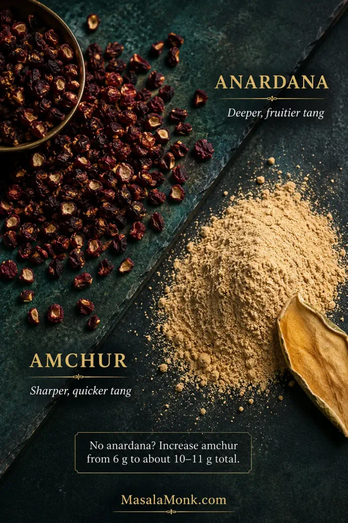 Small dried burgundy anardana seeds beside pale amchur powder on a dark green surface, with a piece of dried mango nearby.