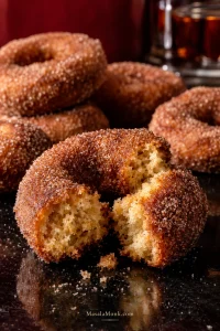 Broken cinnamon-sugar apple cider donut showing a moist cake crumb, with several whole donuts behind it.