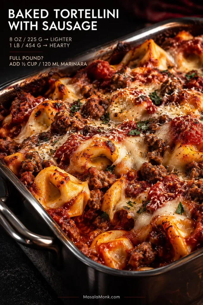 Baked tortellini in marinara with browned pieces of sausage, melted mozzarella and Parmesan in a metal baking dish.